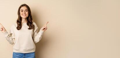Cheerful caucasian woman showing directions, two ways, pointing sideways at variants, choices for customer, standing over beige background photo