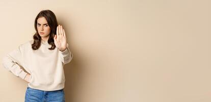 Stop. Serious and confident woman showing extended arm palm, prohibit, forbid smth, blocking something, standing over beige background photo