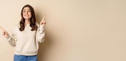 Cheerful caucasian woman showing directions, two ways, pointing sideways at variants, choices for customer, standing over beige background photo