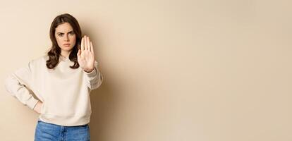 Stop. Serious and confident woman showing extended arm palm, prohibit, forbid smth, blocking something, standing over beige background photo