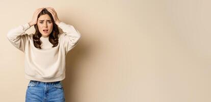 Frustrated and troubled woman facing disaster, holding hands on head in panic, looking anxious, standing over beige background photo