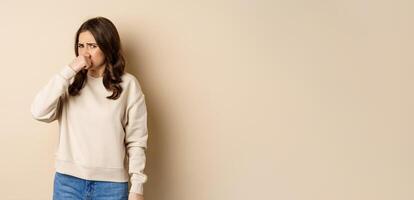 Woman expressing disgust of bad disgusting smell, close nose and grimaing from dislike, standing over beige background photo