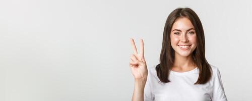 Portrait of beautiful and happy brunette girl smiling and showing peace gesture, standing white background photo
