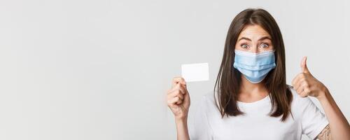 Covid-19, health and social distancing concept. Close-up of pretty brunette girl in medical mask, showing credit card and thumbs-up in approval photo
