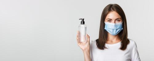 Covid-19, health and social distancing concept. Close-up of attractive smiling girl in medical mask, showing hand sanitizer, recommend antiseptic photo
