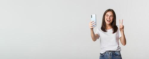 Attractive brunette girl looking happy and taking selfie on smartphone, showing peace gesture photo