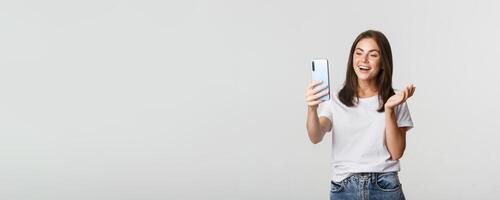 Friendly attractive girl calling friend, smiling and having conversation, holding smartphone, white background photo
