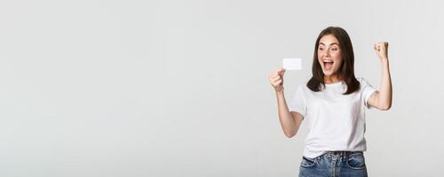 Joyful good-looking girl rejoicing and looking at credit card, fist pump while triumphing, white background photo