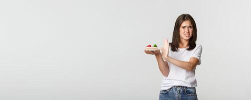 Disgusted young woman rejecting eating awful cake, making refusal gesture and grimacing displeased photo