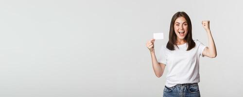 Rejoicing beautiful girl showing credit card and triumphing, fist pump happy, white background photo