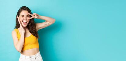 Excited girl screaming from joy, ready for summer holidays, showing v-sign and smiling happy, standing happy against blue background photo