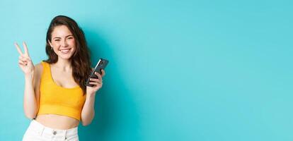 Technology and lifestyle concept. Beautiful cheerful girl sending positive vibes, smiling and showing peace sign, using cellphone, standing against blue background photo