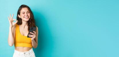 Technology and lifestyle concept. Beautiful girl with happy smile, showing okay sign in approval, say yes, holding smartphone, standing against blue background photo