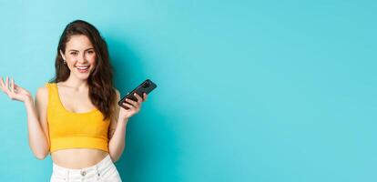 elegante contento mujer participación teléfono inteligente, riendo y sonriente seguro de sí mismo, chateando en móvil teléfono, en pie terminado azul antecedentes foto