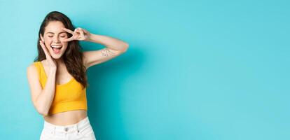 Excited girl screaming from joy, ready for summer holidays, showing v-sign and smiling happy, standing happy against blue background photo