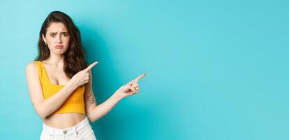 Portrait of sad and gloomy girl sulking with crying face, pointing fingers right at something scary, standing worried against blue background photo
