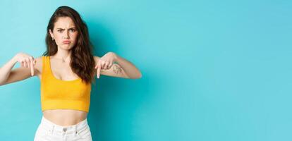 Confused frowning woman looking displeased, need explanation, pointing fingers down at something strange or unpleasant, standing over blue background photo