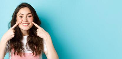 Portrait of happy young woman showing her clean glowing skin and blush on cheeks, pointing fingers at face, smiling satisfied at camera, blue background photo