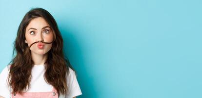 Close up portrait of funny silly girl playing, making squinting face and moustache with hair strand above lips, standing over blue background photo