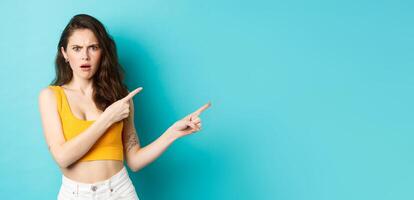Summer lifestyle concept. Confused young woman pointing at direction right, showing something strange, frowning and looking shocked at camera, standing over blue background photo