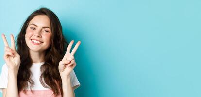 Close up portrait of attractive positive girl saying cheeze, smiling and showing v-signs peace, standing over blue background photo