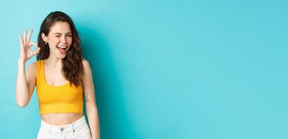 Young cheerful lady in summer clothes winking and saying yes, alright gesture, making OK sign in approval, standing against blue background photo