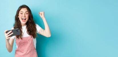 Excited beautful girl winning on mobile phone, holding smartphone and shouting yes with joyful face and fist pump, smiling amazed at camera, blue background photo