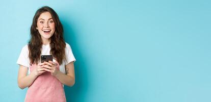 Portrait of excited brunette girl looking amazed at camera while using smartphone, holding mobile phone and smiling cheerful, standing against blue background photo