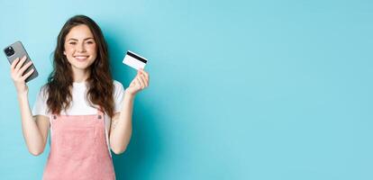 Smiling cute girl showing plastic credit card and smartphone, order something in store via mobile app, shop online, or pay for order, standing against blue background photo