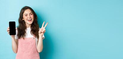 Cute young woman smiling and showing v-sign with empty smartphone screen, demonstrate app or mobile store, standing against blue background photo