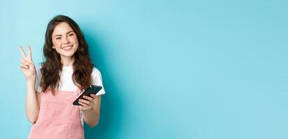 Online shopping. Cute and cheerful young woman smiling, holding smartphone and showing peace v-sign at camera, standing against blue background photo