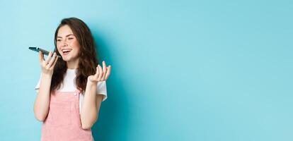 Portrait of smiling woman holding phone near lips and talking, using app translator on smartphone or recording voice message, speaking with speakerphone, standing over blue background photo