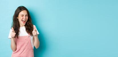 You can do this. Beautiful cheerful brunette girl winking and smiling, pointing fingers at camera, praising good job, inviting to event, standing against blue background photo