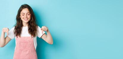 Cute caucasian girl in dungarees pointing and looking down, smiling joyful, showing advertisement on bottom copy space, standing over blue background photo