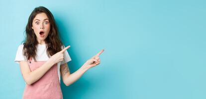 Excited and surprised young woman open mouth, say wow and pointing fingers right at logo, staring impressed at camera while talking about new product or news, blue background photo