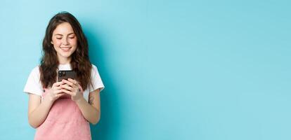Portrait of young woman using smartphone, reading message or chatting in app, standing over blue background and smiling happy photo