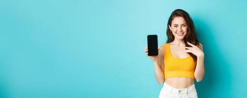 Stylish caucasian woman in summer outfit showing empty smartphone screen and smiling, demonstrate an app or online store, standing over blue background photo