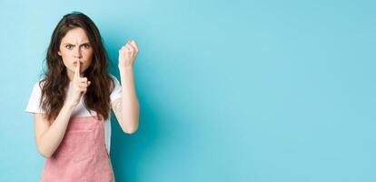 Serious and angry brunette girl shaking fist and frowning with disapproval, scolding person bad behaviour, telling to keep quiet, holding finger on lips, blue background photo