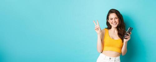 Technology and lifestyle concept. Beautiful cheerful girl sending positive vibes, smiling and showing peace sign, using cellphone, standing against blue background photo
