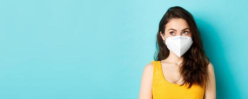 Covid-19, coronavirus and social distancing. Close up portrait of young modern woman in respirator, looking at upper left corner logo, standing against blue background photo