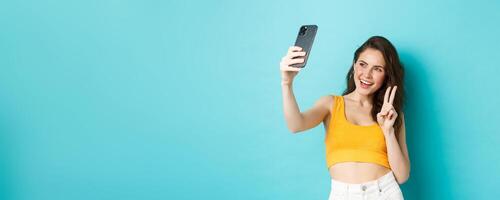 Technology and lifestyle concept. Stylish young woman taking selfie in her summer clothes, showing v-sign at smartphone camera, standing over blue background photo