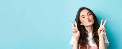 Close up portrait of glamour brunette girl making kissing face, showing kawaii v-sign and smiling cute at camera, standing happy against blue background photo
