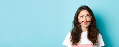 Close up portrait of funny silly girl playing, making squinting face and moustache with hair strand above lips, standing over blue background photo