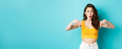 Young curious female customer asking question about item, pointing fingers down with questioned face, standing against blue background photo