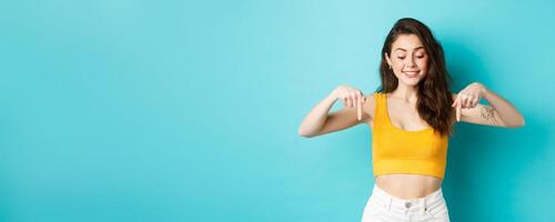 Portrait of stylish natural girl in summer crop top, pointing and looking down with dreamy smile, seeing something interesting on bottom advertisement, blue background photo