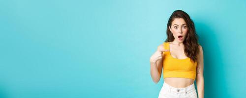 Portrait of young confused woman pointing at herself, looking with surprise and disbelief at camera, being accused or chosen unexpectedly, standing over blue background photo