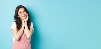 Portrait of beautiful girl dreaming, looking aside at logo banner and thinking, standing romantic against blue background, smiling and imaging something cute, blue background photo