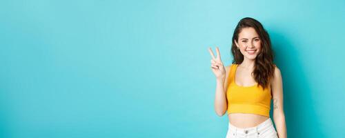 Summer and lifestyle concept. Beautiful girl tourist in crop top, posing with v-sign and smiling happy at camera, standing against blue background photo