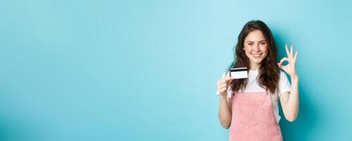 Beautiful sassy girl recommending plastic credit card, showing okay sign and looking confident at camera, smiling satisfied, standing over blue background photo