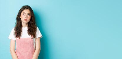 Portrait of sad and gloomy young brunette girl with glamour make up, looking at upper left corner upset and thoughtful, feeling moody, standing against blue background photo